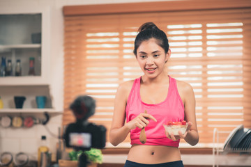Fototapeta na wymiar Asian blogger woman make vlog how to diet and lost weight, Young female using camera recording when she eating yogurt in the kitchen. Lifestyle influencer women healthy concept.
