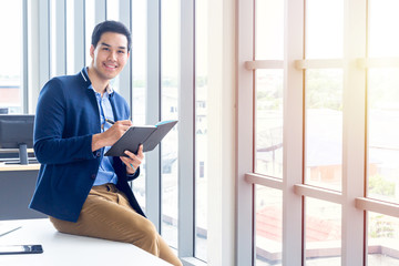 A businessman wearing a suit and a long-sleeve shirt inside and sitting smart poses in a modern office. In his hand have a pen and a notebook in action reading or writing something.