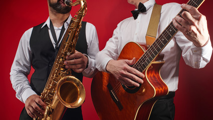 Group of two musicians, male jazz band, guitarist and saxophonist in classical costumes improvise on musical instruments in a studio on  red background