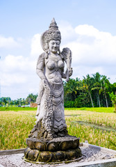 An ancient statue of a woman in a traditional Balinese style against a field and jungle background. Bali, Indonesia