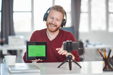 Handsome Caucasian employee having presentation over the internet. In hands is tablet and on head are headphones. Start up business concept.