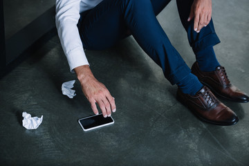 cropped view of businessman sitting on floor near smartphone with blank screen