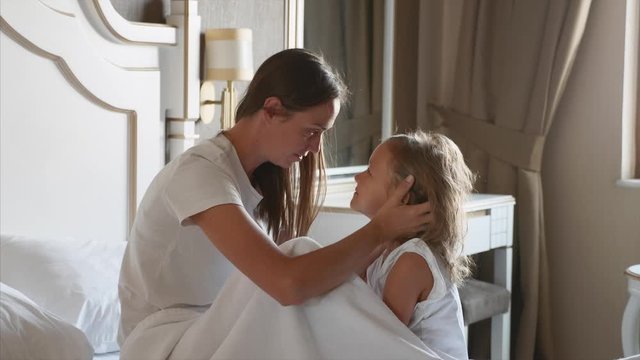 Young mother caresses her little daughter in bedroom at home. They sits on the bed together face to face, mom strokes child and kisses her.