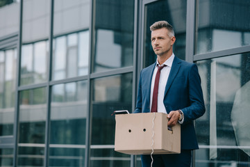 dismissed businessman in suit standing near building with carton box