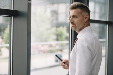 Fototapeta premium handsome man holding smartphone with blank screen near window