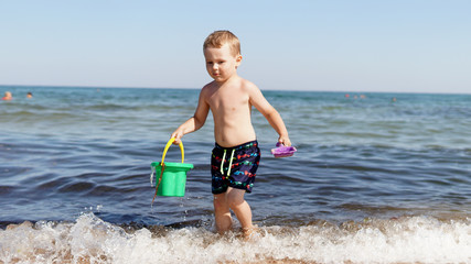 Little boy walking on sea beach along the sea wave and holds a pail in his hands