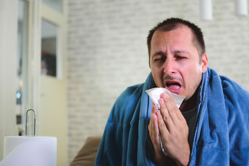 Young man holding his throat in pain and drinking healthy herbal tea. Portrait of sick men covered with blue blanket. - obrazy, fototapety, plakaty