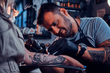 Fototapeta na wymiar Dilligent focused tattoo artist is creating new tattoo on young woman's hand at tatoo studio.