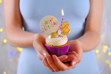 Woman holding happy birthday cupcake with burning candle in purple wrap, cream cheese frosting swirl and golden chocolate decoration on white wooden textured table. Close up, copy space, background.