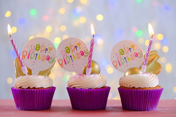 Sweet organic happy birthday cupcake with burning candle in purple wrap, cream cheese frosting swirl and golden chocolate decoration on white wooden textured table. Close up, copy space, background.