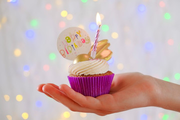 Sweet organic happy birthday cupcake with burning candle in purple wrap, cream cheese frosting swirl and golden chocolate decoration on white wooden textured table. Close up, copy space, background.