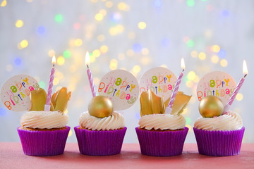 Sweet organic happy birthday cupcake with burning candle in purple wrap, cream cheese frosting swirl and golden chocolate decoration on white wooden textured table. Close up, copy space, background.