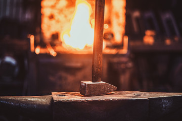 Hammer on anvil at dark blacksmith workshop with fire in stove at background.
