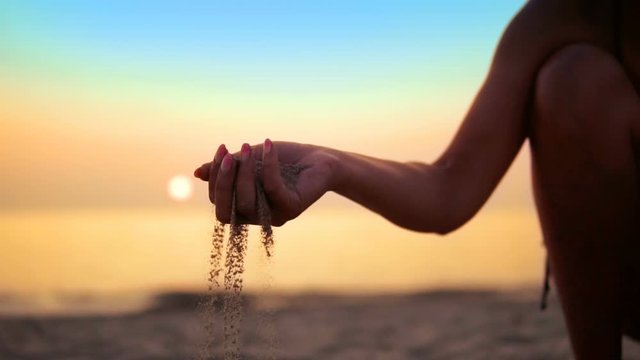 Close-up of female hand releasing dropping sand against sea sunset horizon. Sand flowing through the hands against orange sky. Summer beach holiday vacation concept, SLOW MOTION