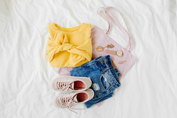 Yellow sweater, blue jeans, eco bag and sneakers on bed on white sheet. Women's stylish autumn or winter outfit. Trendy  clothes collage. Flat lay, top view.
