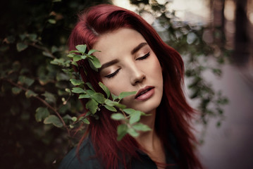 Young fashionable woman in romantic green dress and red hair, posing near houses in park.