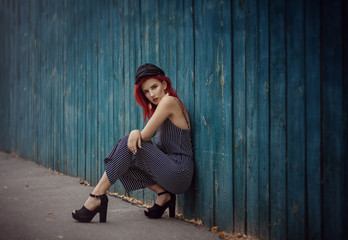 Young fashionable woman in clothes in a strip and red hair, posing on a blue background on the street.