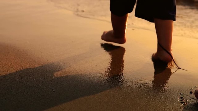 Following and undefined baby feet walking on beach at sunset with sea waves splashing his barefoot legs, SLOW MOTION