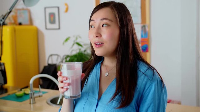 Alluring pensive Asian young woman drinking tasty fruit smoothie with coco milk at kitchen, cute smiling female in blue shirt with long brunette hair enjoying fresh yellow fridge on background