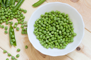 White bowl with green snap sugar peas. Flat lay. Peeled beans scattered on wooden table, single open pod and few whole pods.