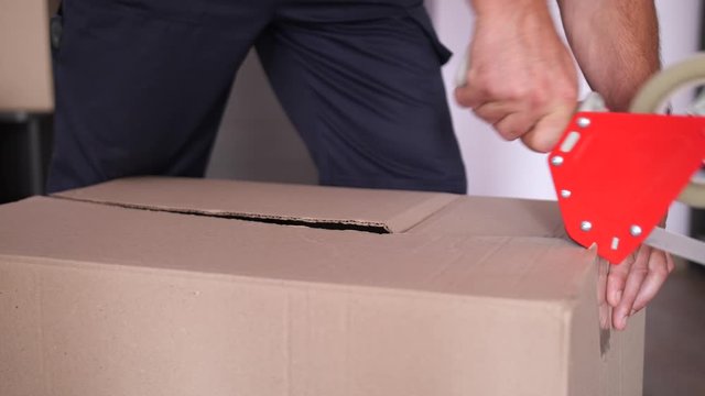 Close-up of male hands packing removal cardboard box with adhesive tape using applicator. Employee of relocation service sealing package with scotch tape while packing client's things during moving