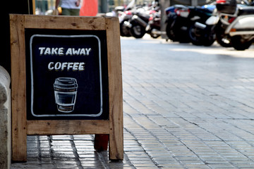 Old-fashion sidewalk sandwich board sign on the pavement with the message Take Away Coffee. Unidentifiable people in the background