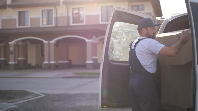 Professional worker of delivery service in cap and overalls taking cardboard package out from van and carrying to customer. Adult bearded employee, working in home delivery, unloading car trunk