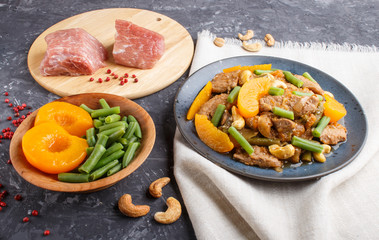 Fried pork with peaches, cashew and green beans on a black concrete background, close up. raw meat, red pepper, linen textile.