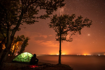 The man are sitting on the lake, looking at the starry sky. Small tent and tree on the shore. Night landscape.