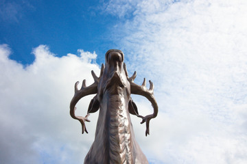 A large deer is made of wood against the sky. The deer has large horns that, against the sky, seem to touch the clouds. The head of a deer is raised to the sky.