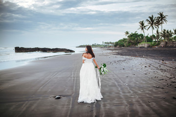 Fototapeta na wymiar Bride on a beach in the blue water with a bouquet in hands on sunset