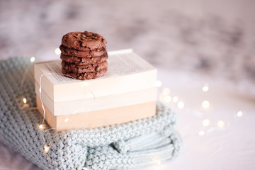 Stack of tasty chocolate cookies on open books and knitted sweater over Christmas lights closeup. Good morning. Winter holiday season. Homemade bakery indoors. New Year.
