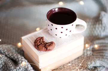 Cup of tea with chocolate cookie with bite on open books over Christmas lights close up. Winter holiday season. Good morning. New Year.