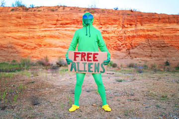 a man dressed in a green alien carnival costume suit at a lone rally with a sign "free aliens" on the background of a desert canyon