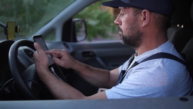 Worker of delivery service in uniform using cellphone navigator app setting route to client while sitting in car. Adult male in cap and overalls using smartphone to check route before delivering order