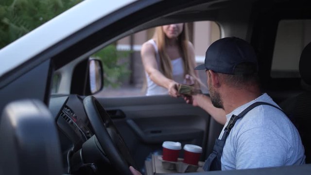 Delivery man in uniform sitting in van giving ordered cups of coffee drink to attractive woman customer. Blonde female client taking order from food delivery courier through car window and giving tips