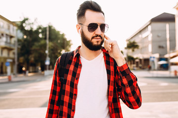 Stylish guy with a beard in wireless headphones walks around the city and listens to music.