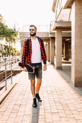 Stylish guy with a beard in wireless headphones walks with a skateboard around the city