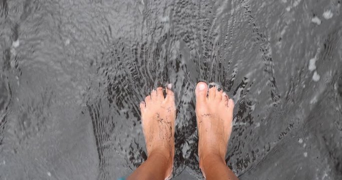 Beach feet closeup - barefoot woman walking in ocean water waves on black sand beach. Female young adult legs and toes relaxing on summer vacation travel. From Hawaii, USA
