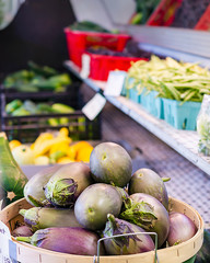 Bushel of egg plant at a roadside stand selling fresh produce.  Additional vegetables in the background, including beans, yellow squash, and cucumbers.