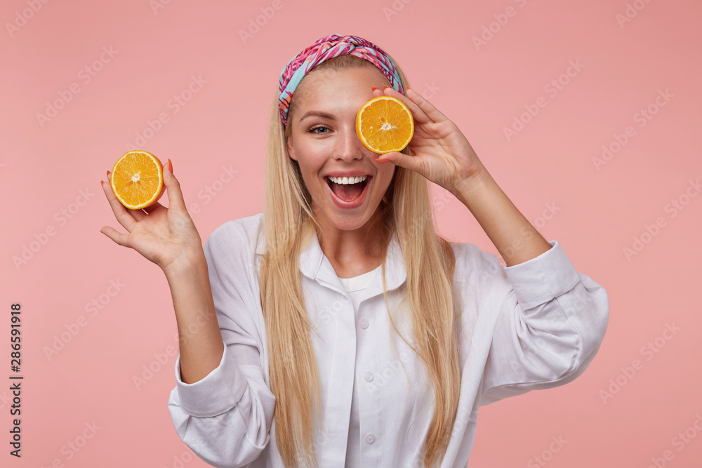 Wall mural Attractive young woman with broad charming smile making fun over pink background, holding cut orange near her eye and fooling on camera