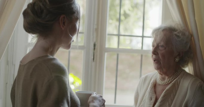 Multigeneration women talking together. Senior grandma woman smiling with her granddaughter visiting near window drinking tea or coffee.White hair elderly grandmother at home.4k slow motion