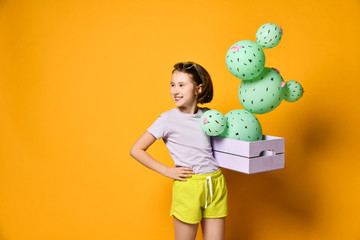 Young brunette girl hold cactus balloon in pastel pink wooden box. Flowers are made of balloons with pink flowers and painted spines. Creative idea minimal concept on yellow background