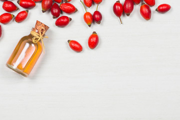 Glass bottle of rosehip seed essential oil with fresh rose hip fruits on white rustic background. Dogrose oil with fresh dog roses