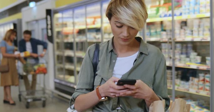 Camera zooming in on the Caucasian pretty and stylish young woman with short blond hair tapping and texting on the smartphone while shopping in the supermarket.