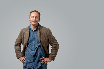 smiling old casual man in leather jacket with hands in pockets standing against gray studio wall