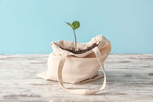Eco bag with soil and plant on wooden table against color background