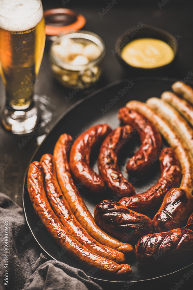 Wall mural Octoberfest dinner table concept with grilled veal and pork sausages, sauce in jar, pickled vaggies and beer in glass over dark background, selective focus, close-up