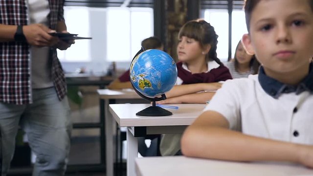 Male teacher of elementary school walking between desks puts computers tablet on desk pupils for learning