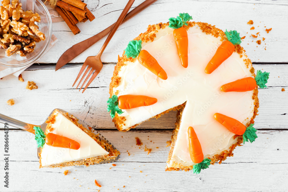 Poster Homemade carrot cake with cream cheese frosting. Above view table scene  with slice being removed. Close up over a white wood background.